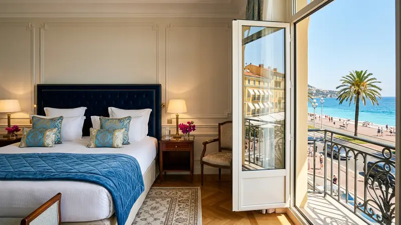 Chambre d'hôtel niçois haut de gamme avec literie blanche immaculée, fenêtre donnant sur la Promenade des Anglais et la Méditerranée, lumière dorée d'été