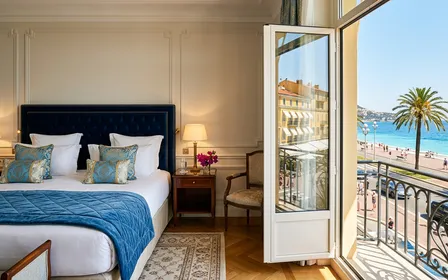 Chambre d'hôtel niçois haut de gamme avec literie blanche immaculée, fenêtre donnant sur la Promenade des Anglais et la Méditerranée, lumière dorée d'été