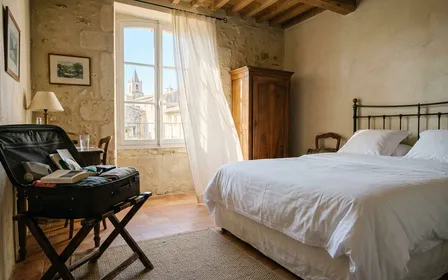 Chambre d'hôtel française avec valise ouverte sur un porte-bagage, lit blanc en second plan, lumière naturelle de matin d'été filtrant à travers un voilage clair, ambiance de premier jour de vacances