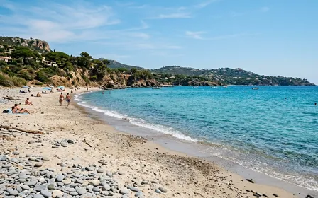 Plage française méditerranéenne en fin de matinée avec sable clair, mer bleu turquoise en second plan et quelques galets au premier plan, ambiance estivale calme sans foule