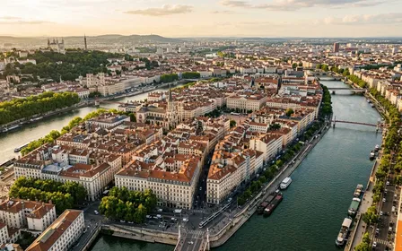 Vue aérienne d'une grande ville française avec toits, parcs urbains et rives fluviales sous une lumière dorée de fin d'après-midi d'été, ambiance de métropole en juillet