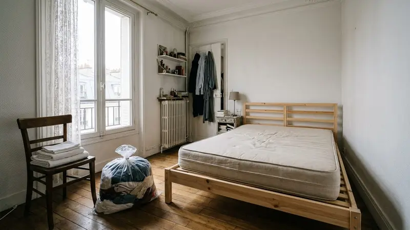 Chambre française avec matelas nu posé sur un sommier en bois clair, quelques draps blancs pliés sur une chaise et vêtements dans un sac en plastique fermé, ambiance de préparation avant traitement, lumière naturelle de matinée
