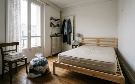 Chambre française avec matelas nu posé sur un sommier en bois clair, quelques draps blancs pliés sur une chaise et vêtements dans un sac en plastique fermé, ambiance de préparation avant traitement, lumière naturelle de matinée