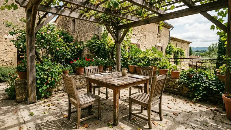 Coin de terrasse française d'été abandonnée avec une table en bois et des chaises vides sous une pergola, ambiance de mi-journée chaude avec quelques feuilles au sol, sans convives visibles