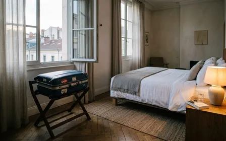 Valise de voyage rigide posée sur un porte-bagage en hauteur dans une chambre d'hôtel française claire, avec lit fait au fond et lampe de chevet allumée, ambiance sobre calme de fin de journée