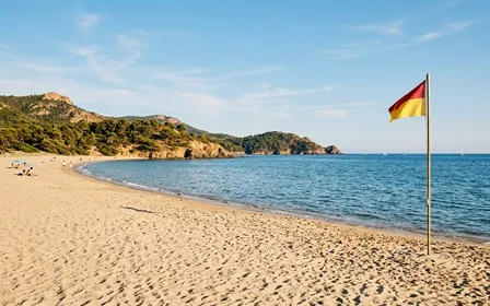 Plage française du Var en fin d'été avec sable clair, mer bleue calme au fond et drapeau de baignade planté dans le sable, ambiance lumineuse d'après-midi ensoleillé sans baigneur