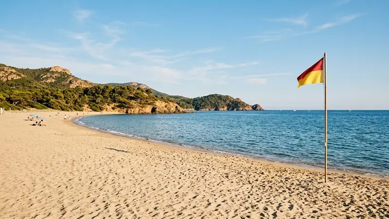 Plage française du Var en fin d'été avec sable clair, mer bleue calme au fond et drapeau de baignade planté dans le sable, ambiance lumineuse d'après-midi ensoleillé sans baigneur