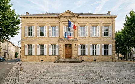 Façade de mairie française classique avec drapeau tricolore visible et parvis en pierre en fin de matinée d'été, ambiance calme documentaire sans passant visible