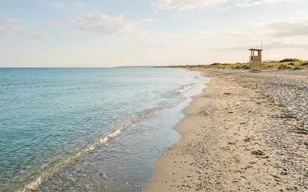 Plage méditerranéenne française de sable et galets clairs en été avec vaguelettes calmes et poste de surveillance en bois au loin, ambiance sobre lumière naturelle de fin d'après-midi documentaire aucun humain visible plan large