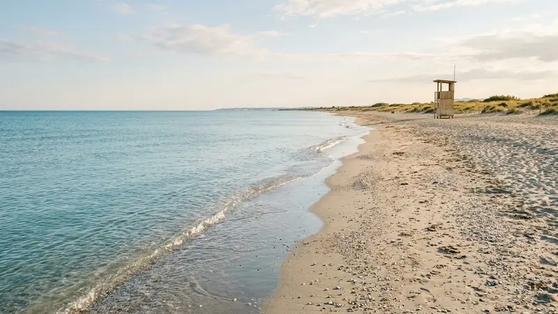 Plage méditerranéenne française de sable et galets clairs en été avec vaguelettes calmes et poste de surveillance en bois au loin, ambiance sobre lumière naturelle de fin d'après-midi documentaire aucun humain visible plan large