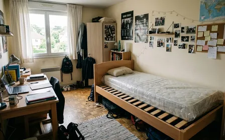 Chambre étudiante française contemporaine avec lit simple à structure en bois clair sommier apparent et matelas nu recouvert de son emballage plastique, ambiance sobre lumière naturelle douce de fin de matinée aucun humain visible plan large documentaire