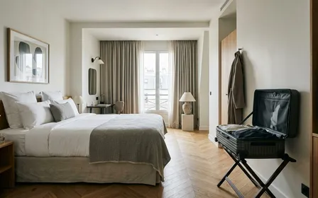 Chambre d'hôtel française contemporaine avec valise rigide posée sur porte-bagages en métal à distance du lit et rideaux tirés au fond, ambiance sobre lumière naturelle douce de fin de matinée aucun humain visible plan large documentaire