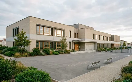 Façade extérieure d'école primaire française contemporaine en brique claire et béton avec cour de récréation vide et arbustes en pied de mur, ambiance sobre lumière naturelle documentaire de fin d'après-midi aucun humain visible plan large