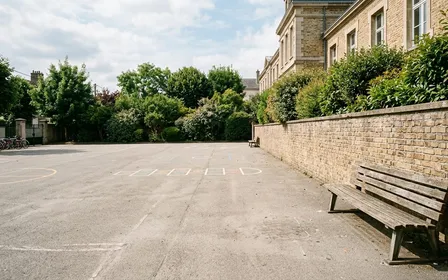 Cour de récréation d'école primaire française avec sol en enrobé gris clair et banc en bois adossé à un mur en brique claire, ambiance sobre lumière naturelle documentaire de fin de matinée aucun humain visible plan large