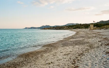 Plage méditerranéenne française de sable et petits galets clairs en fin d'après-midi de fin août avec mer calme et poste de surveillance en bois au loin, ambiance sobre lumière naturelle documentaire aucun humain visible plan large
