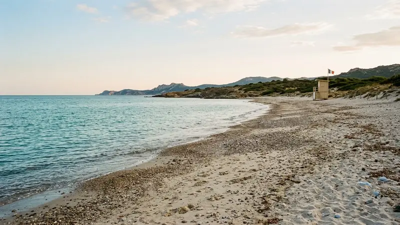 Plage méditerranéenne française de sable et petits galets clairs en fin d'après-midi de fin août avec mer calme et poste de surveillance en bois au loin, ambiance sobre lumière naturelle documentaire aucun humain visible plan large