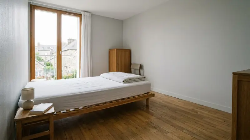 Chambre française d'un logement contemporain avec matelas isolé du mur sur sommier à lattes et housse blanche hermétique tirée jusqu'aux angles, ambiance sobre lumière naturelle documentaire aucun humain visible plan large