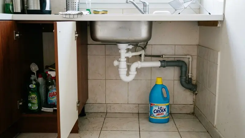 Évier de cuisine française contemporaine avec siphon en plastique blanc visible sous le lavabo et bouteille de javel bleue posée à côté sur le carrelage clair, ambiance sobre lumière naturelle documentaire aucun humain ni insecte visible plan rapproché
