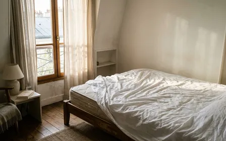 Chambre française contemporaine en début de matinée avec drap blanc froissé sur un matelas nu et lumière naturelle traversant les rideaux entrouverts, ambiance sobre documentaire aucun humain ni insecte visible plan large sur un coin de lit