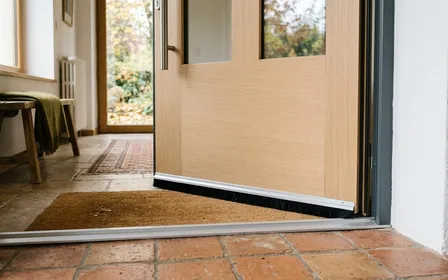 Porte d'entrée française contemporaine en bois clair vue de l'intérieur avec seuil en aluminium propre et joint balai brosse noir visible sous la porte en fin de matinée d'automne, ambiance sobre lumière naturelle documentaire aucun humain ni animal visible plan macro rapproché