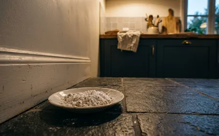 Coin de cuisine française contemporaine en carrelage clair sombre en fin de journée d'automne avec petite soucoupe en porcelaine blanche contenant un mélange de poudre blanche de bicarbonate et sucre glace posée au sol près d'une plinthe, ambiance sobre lumière artificielle douce documentaire aucun humain ni insecte visible plan macro rapproché