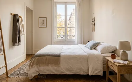 Chambre à coucher française contemporaine d'un logement Airbnb avec lit double soigneusement fait avec draps blancs frais et table de chevet en bois clair en fin de matinée d'automne, ambiance sobre lumière naturelle documentaire aucun humain ni insecte visible plan large horizontal