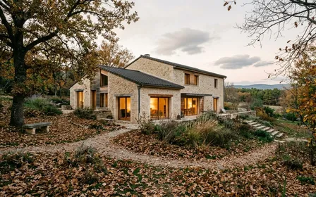 Maison individuelle française contemporaine en pierre claire vue depuis le jardin en fin d'après-midi d'automne avec feuilles mortes brunes au sol et lumière chaude sortant des fenêtres du rez-de-chaussée, ambiance sobre lumière naturelle documentaire aucun humain ni animal visible plan large horizontal