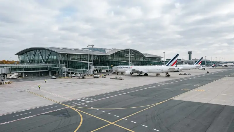 Vue extérieure d'un terminal moderne de l'aéroport Roissy-Charles-de-Gaulle avec avion blanc visible en arrière-plan et tarmac gris propre en fin de matinée d'automne, ambiance sobre lumière naturelle documentaire aucun humain visible plan large horizontal