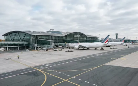 Vue extérieure d'un terminal moderne de l'aéroport Roissy-Charles-de-Gaulle avec avion blanc visible en arrière-plan et tarmac gris propre en fin de matinée d'automne, ambiance sobre lumière naturelle documentaire aucun humain visible plan large horizontal