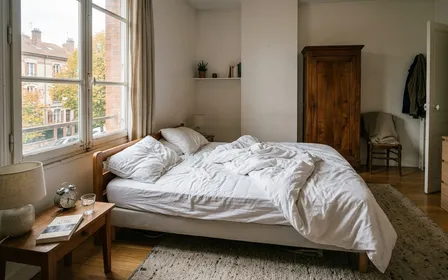 Chambre française contemporaine en début de matinée d'automne avec lit défait et draps blancs relevés révélant le matelas nu blanc en fin de matinée, ambiance sobre lumière naturelle documentaire aucun humain ni animal visible plan large horizontal