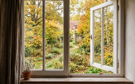 Fenêtre française contemporaine en bois clair peint blanc entrouverte donnant sur un jardin d'automne aux feuilles jaunies en fin de matinée d'octobre, ambiance sobre lumière naturelle documentaire aucun humain ni insecte visible plan large horizontal