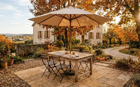 Terrasse française contemporaine en fin d'après-midi ensoleillé d'automne avec table de jardin en bois clair et chaises vides sous un parasol beige clair, ambiance sobre lumière naturelle documentaire aucun humain ni insecte visible plan large horizontal