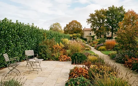 Jardin français contemporain d'automne avec haie de laurier vert dense et coin de terrasse en dalles beige clair en début de matinée, ambiance sobre lumière naturelle documentaire aucun humain ni insecte visible plan large horizontal
