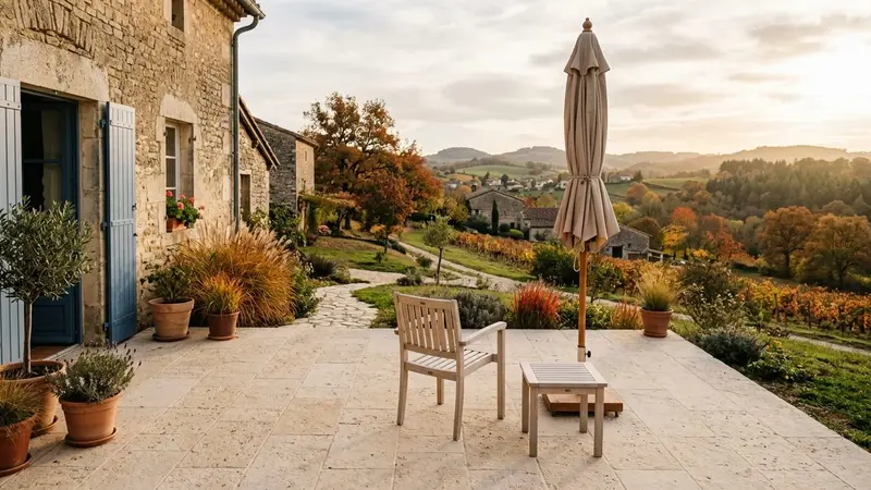 Terrasse française contemporaine en dalles beige clair d'automne avec chaise en bois clair vide et table basse en bois clair sous parasol beige plié en fin d'après-midi, ambiance sobre lumière naturelle documentaire aucun humain ni insecte visible plan large horizontal