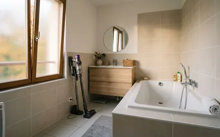 Salle de bains française contemporaine en carrelage clair avec joints silicone blanc propre autour de la baignoire et aspirateur balai posé à côté en début de matinée d'automne, ambiance sobre lumière naturelle documentaire aucun humain ni insecte visible plan large horizontal