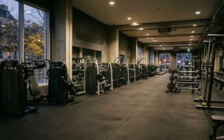 Salle de sport française contemporaine avec machines de musculation alignées et sol en caoutchouc noir en fin d'après-midi d'automne, ambiance sobre lumière artificielle documentaire aucun humain visible plan large horizontal