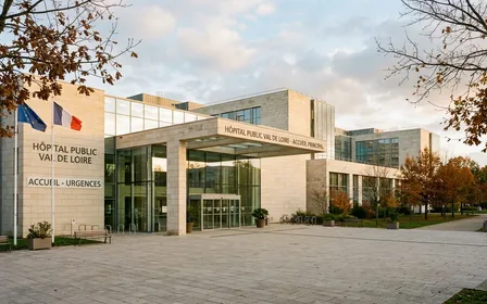 Entrée principale d'hôpital public français contemporain en pierre claire avec parvis dégagé et vitres propres en fin d'après-midi d'automne, ambiance sobre lumière naturelle documentaire aucun humain visible plan large horizontal