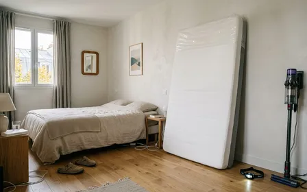 Matelas blanc d'occasion posé verticalement contre un mur clair dans un garage français, lampe torche LED allumée pointée sur une couture latérale, nettoyeur vapeur bleu avec embout buse fine posé au sol à proximité, housse anti-punaises blanche zippée pliée sur une planche à côté, ambiance d'atelier soigné, aucun humain ni animal visible, plan large